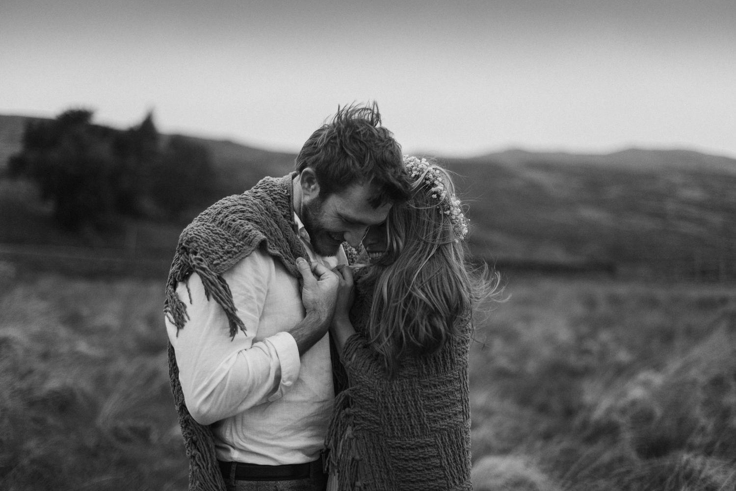 Black and white photo of couple wrapped in a blanket embracing warmly in an open field.