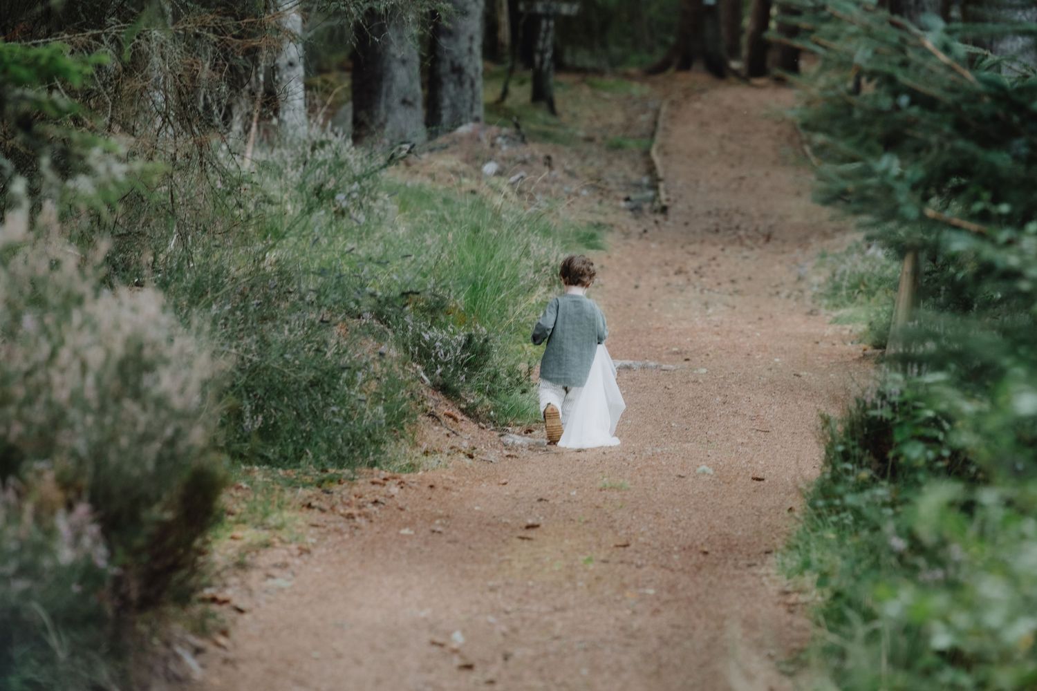 A young child walks alone down a forest trail in a grey outfit during an outdoor woodland wedding.