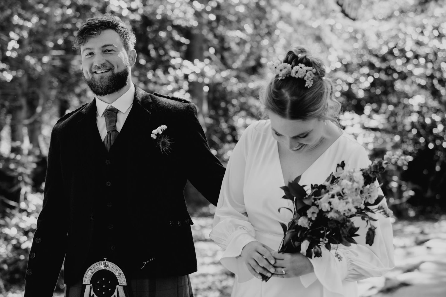 Black and white photo of smiling groom in kilt beside bride holding floral bouquet outdoors.