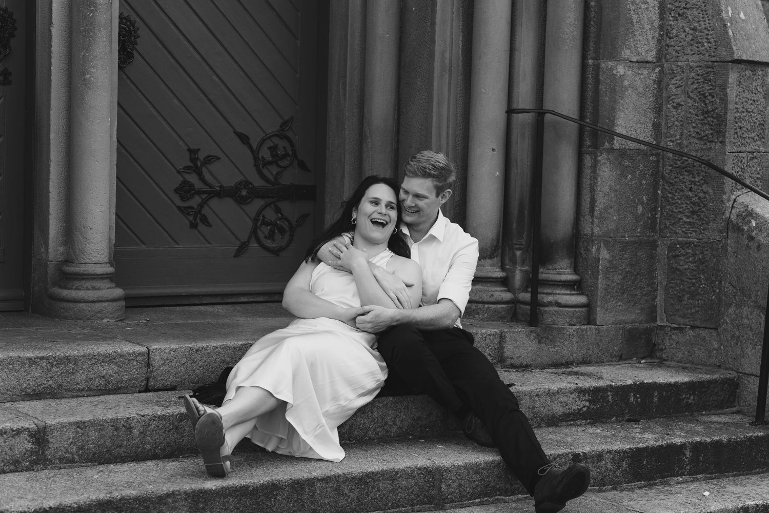 Laughing couple sitting together on stone church steps with large wooden doors in the background.