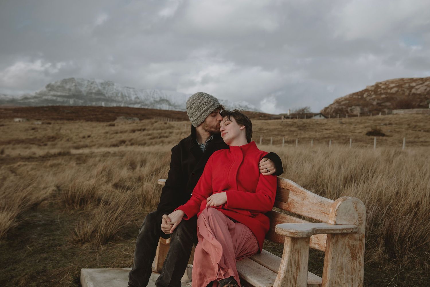 Couple sharing tender moment on rustic wooden bench in moody Scottish moorland setting.