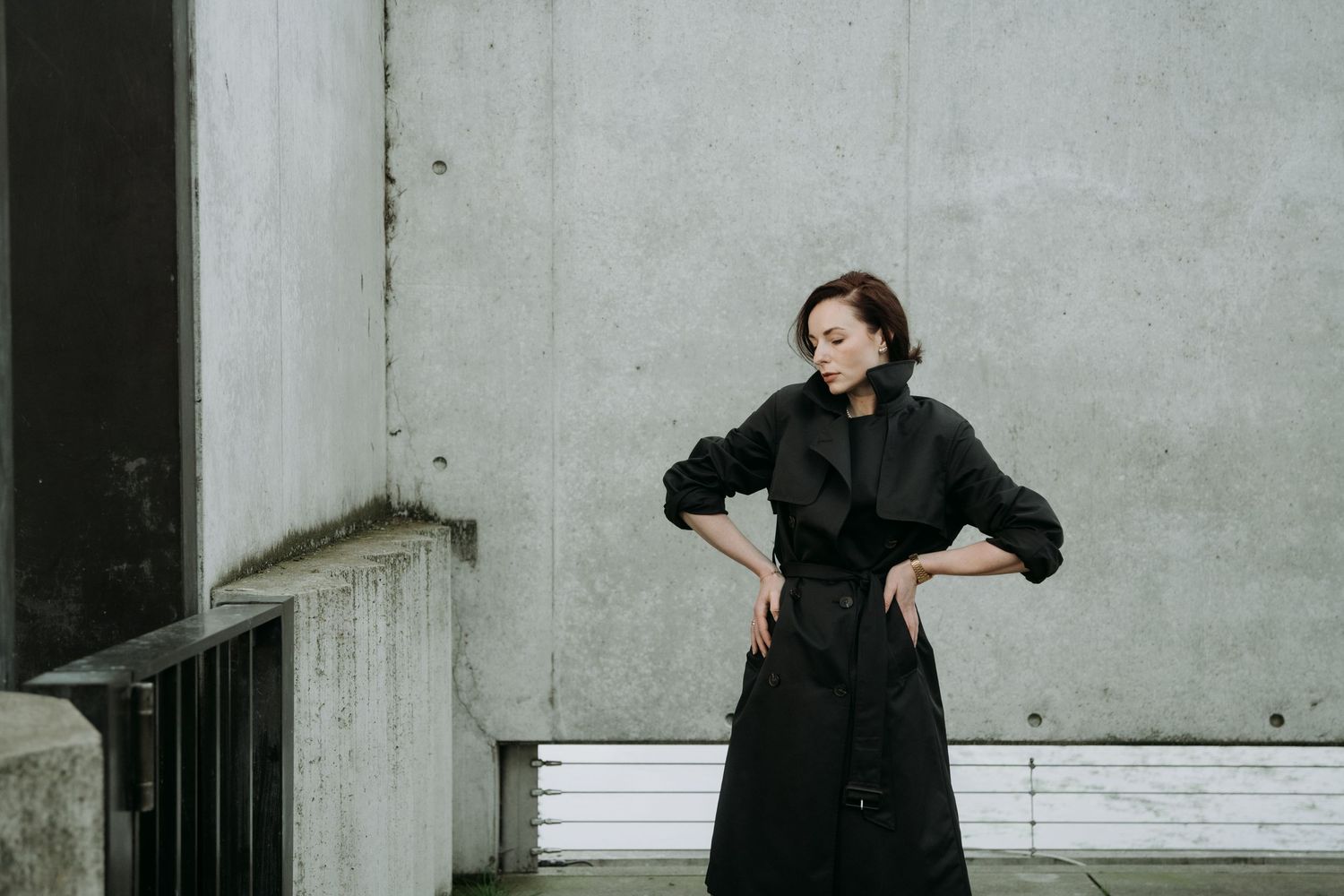 Woman in black oversized coat poses dynamically against a concrete wall in urban setting.