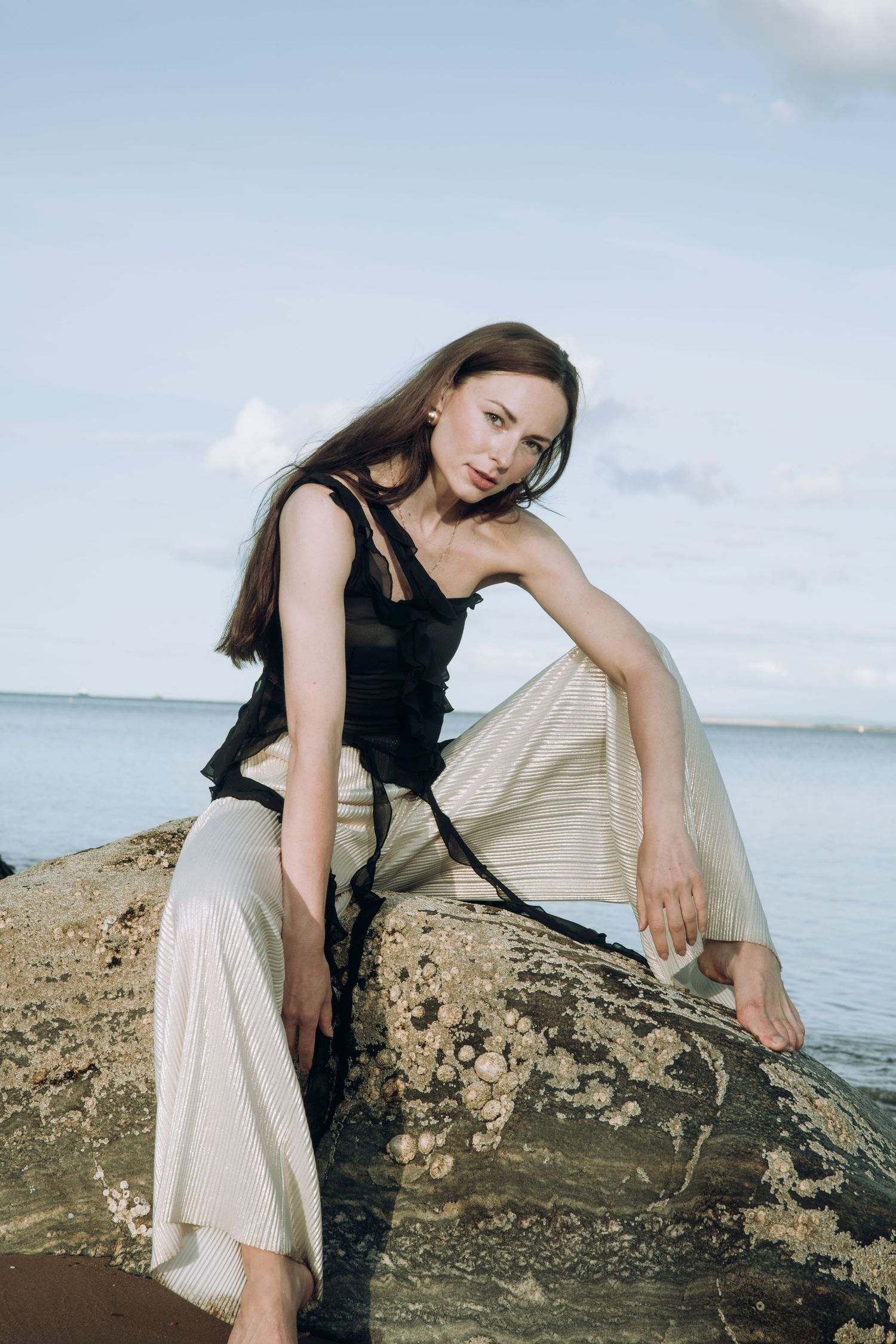 Woman in black top and silver skirt sitting on coastal rocks with ocean in background.