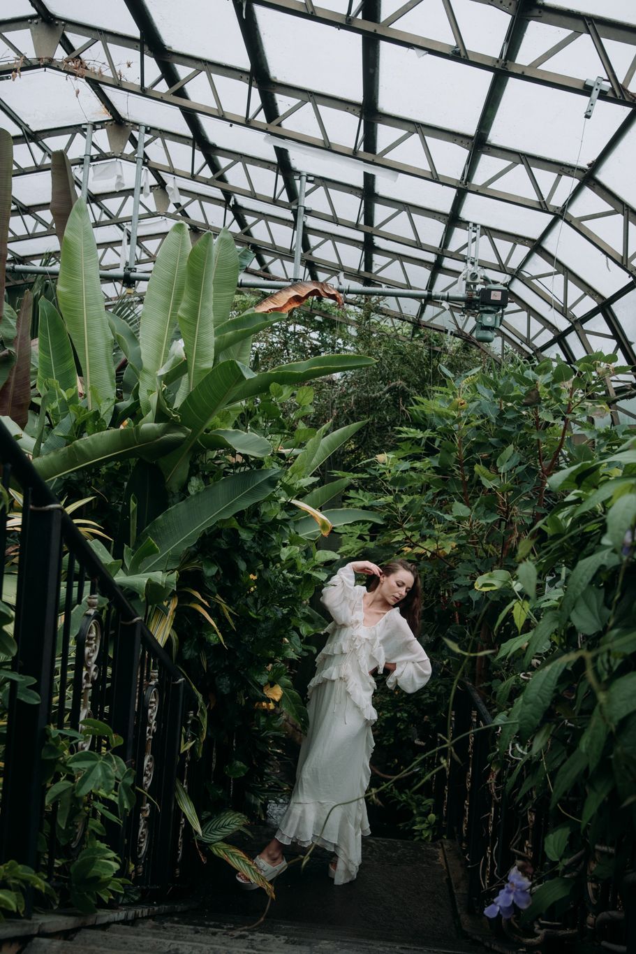 Woman in white ruffled dress walking through a lush tropical glass greenhouse corridor with banana plants.