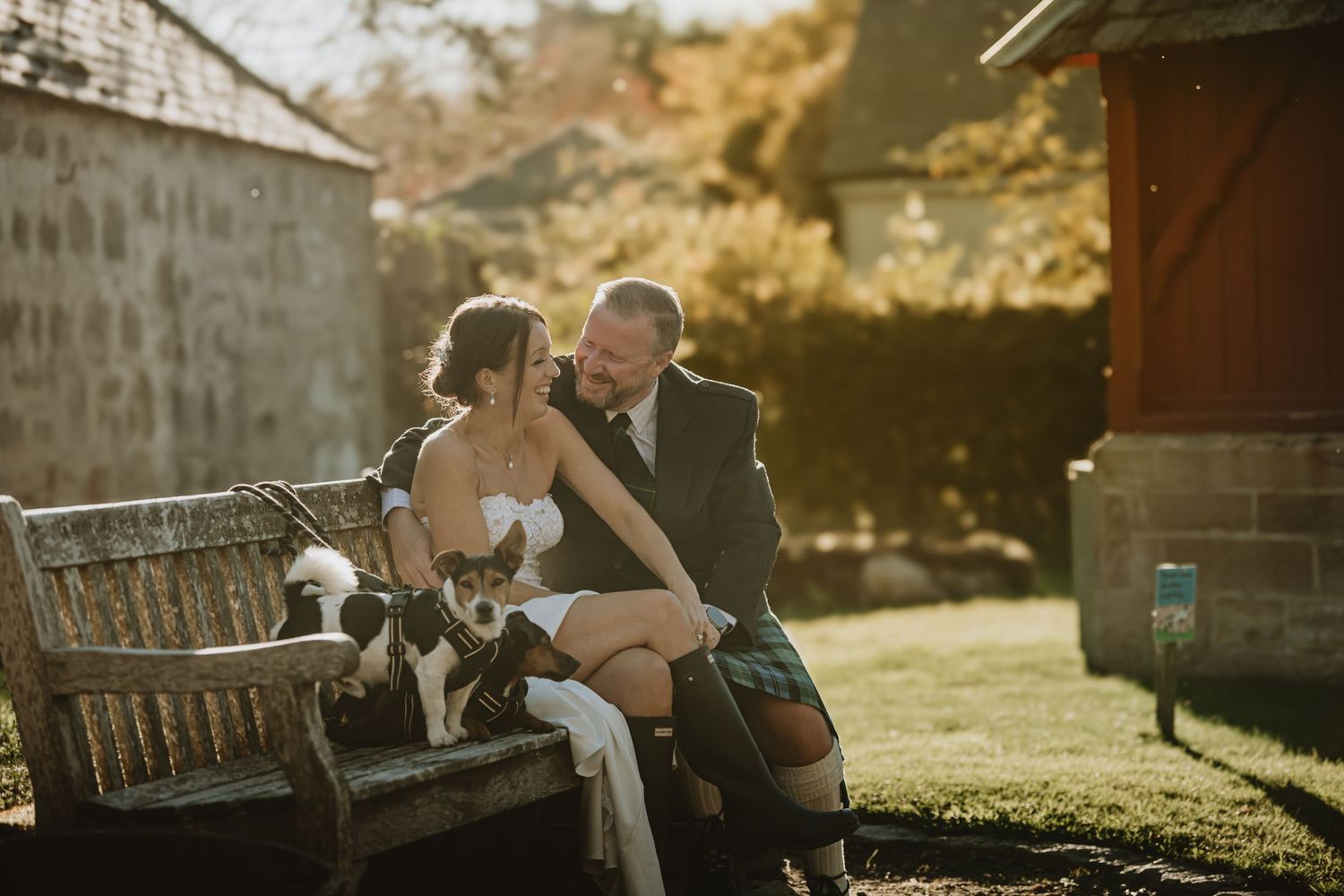 Bride and groom share a tender moment on a garden bench with their dog on a sunny wedding day.