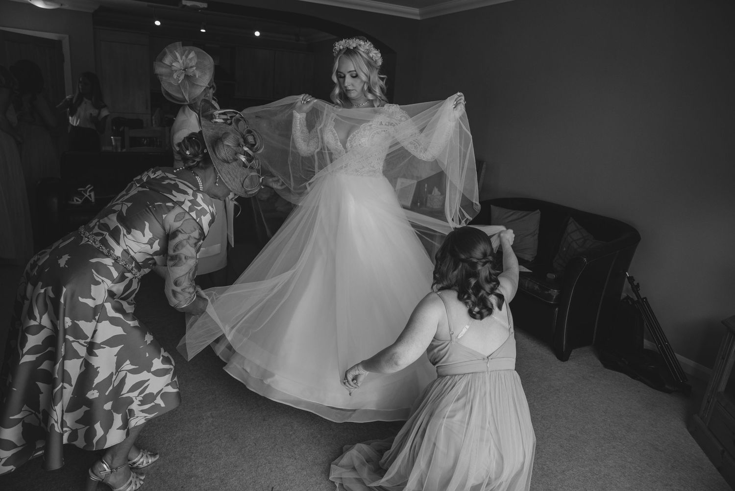 Black and white photo of bridesmaids helping bride arrange her flowing tulle wedding gown and veil.