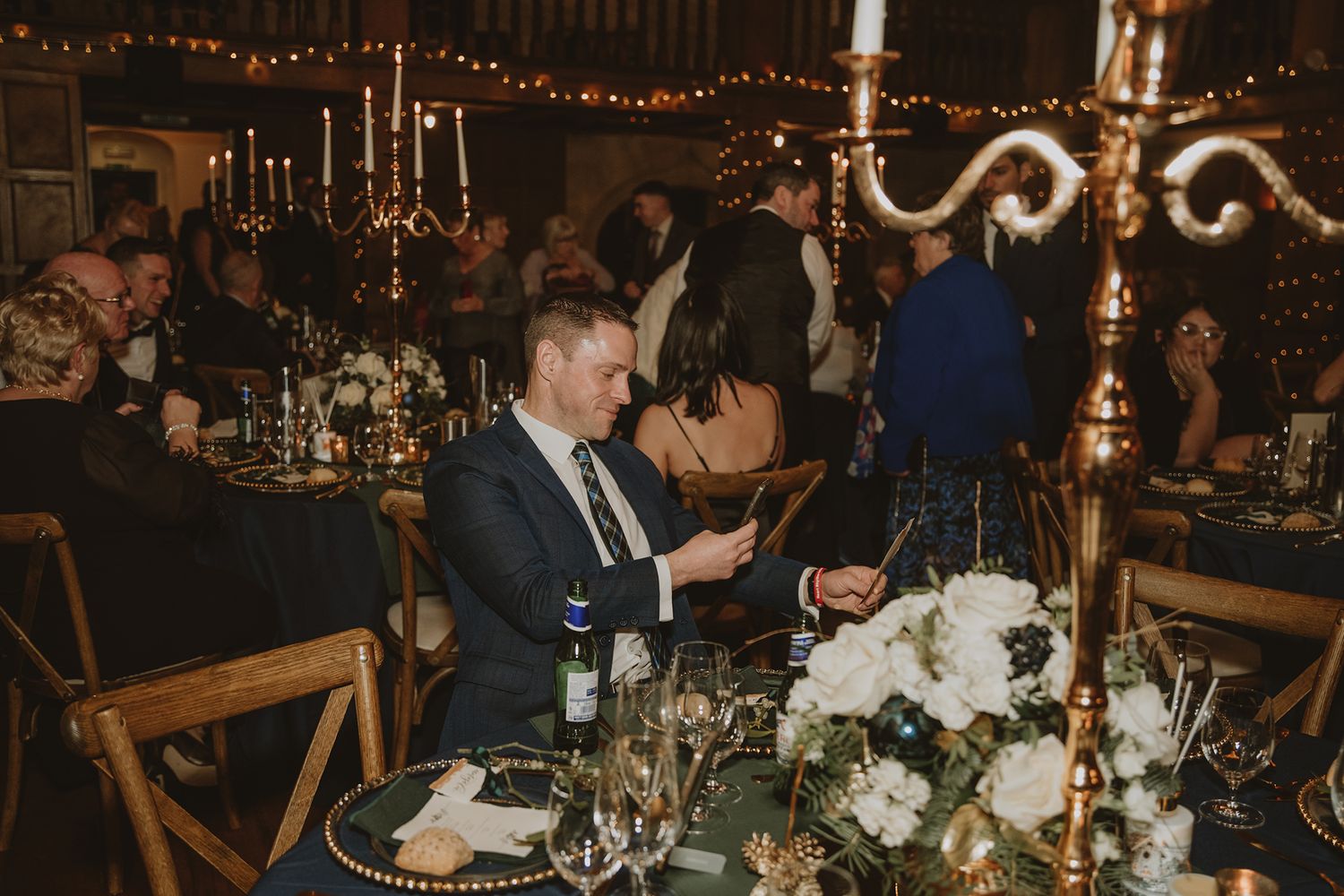 Elegant wedding reception in a barn venue with fairy lights, guests seated at tables with white floral centerpieces and gold candelabras.