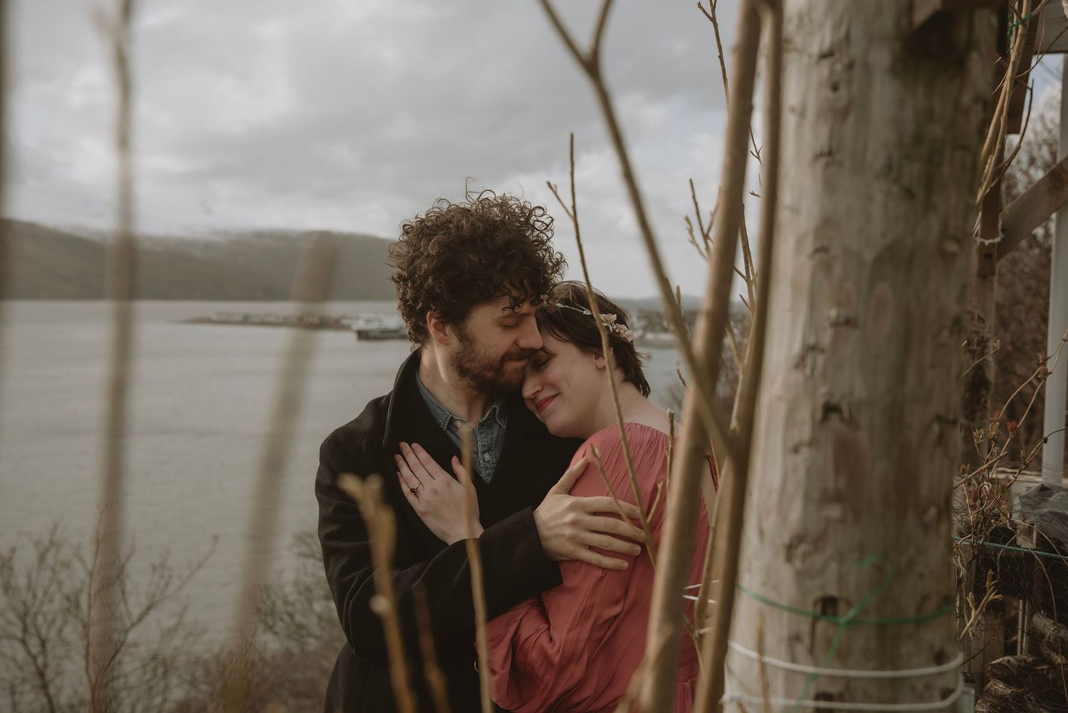 Couple embracing tenderly near a tree by a river on a cloudy day, romantic outdoor engagement photo.