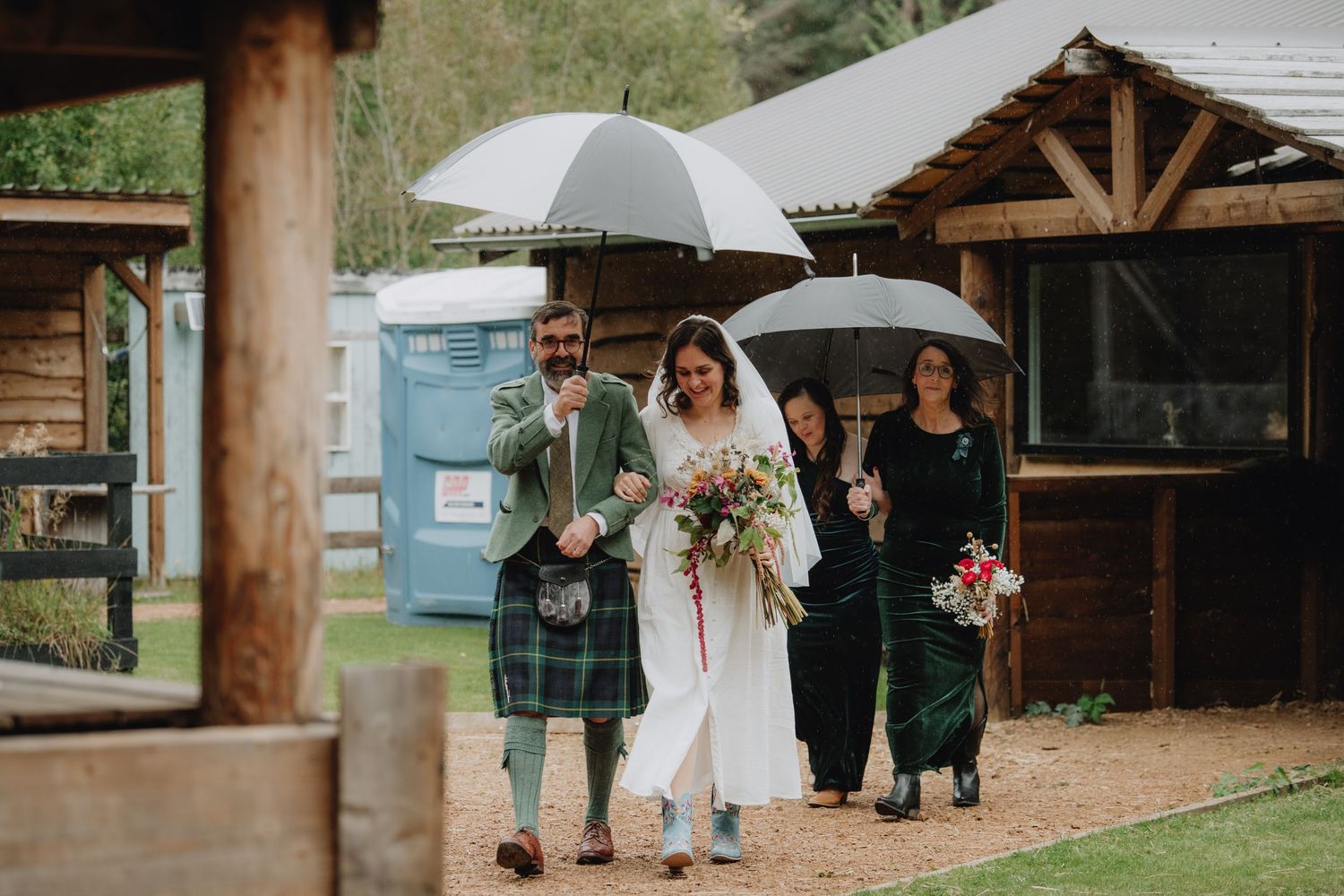 Bride and groom in tartan kilt walking with wedding party under umbrellas on a rainy outdoor wedding day.