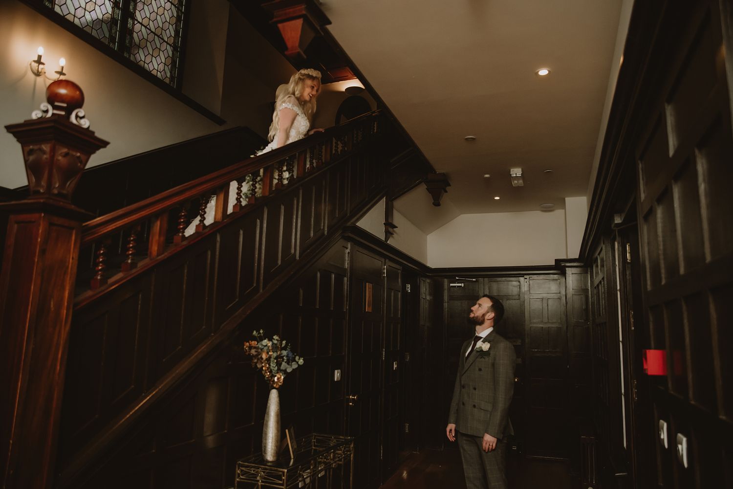 Bride at top of grand wooden staircase looks down at groom standing below in elegant dark-paneled wedding venue.