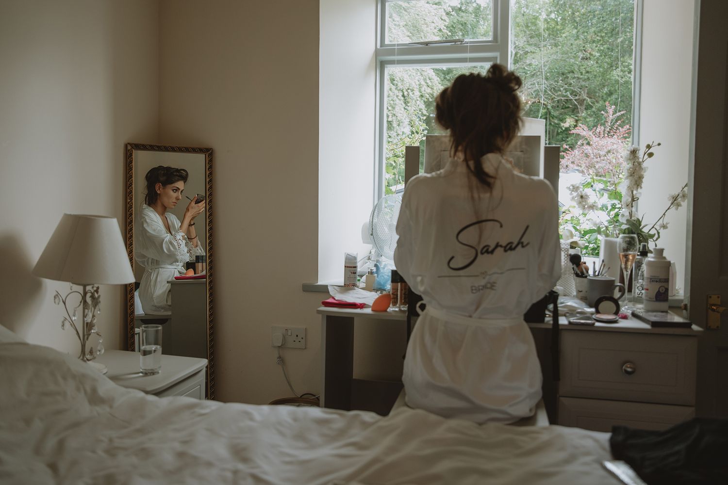 Woman in personalized white bridal robe sitting at vanity mirror getting ready on wedding day.