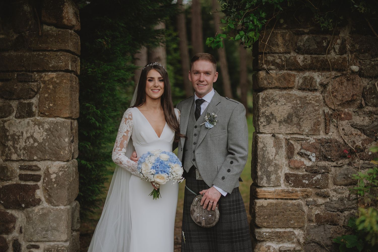 Bride and groom posing between stone pillars, she holds blue bouquet, he wears traditional Scottish kilt and grey jacket.