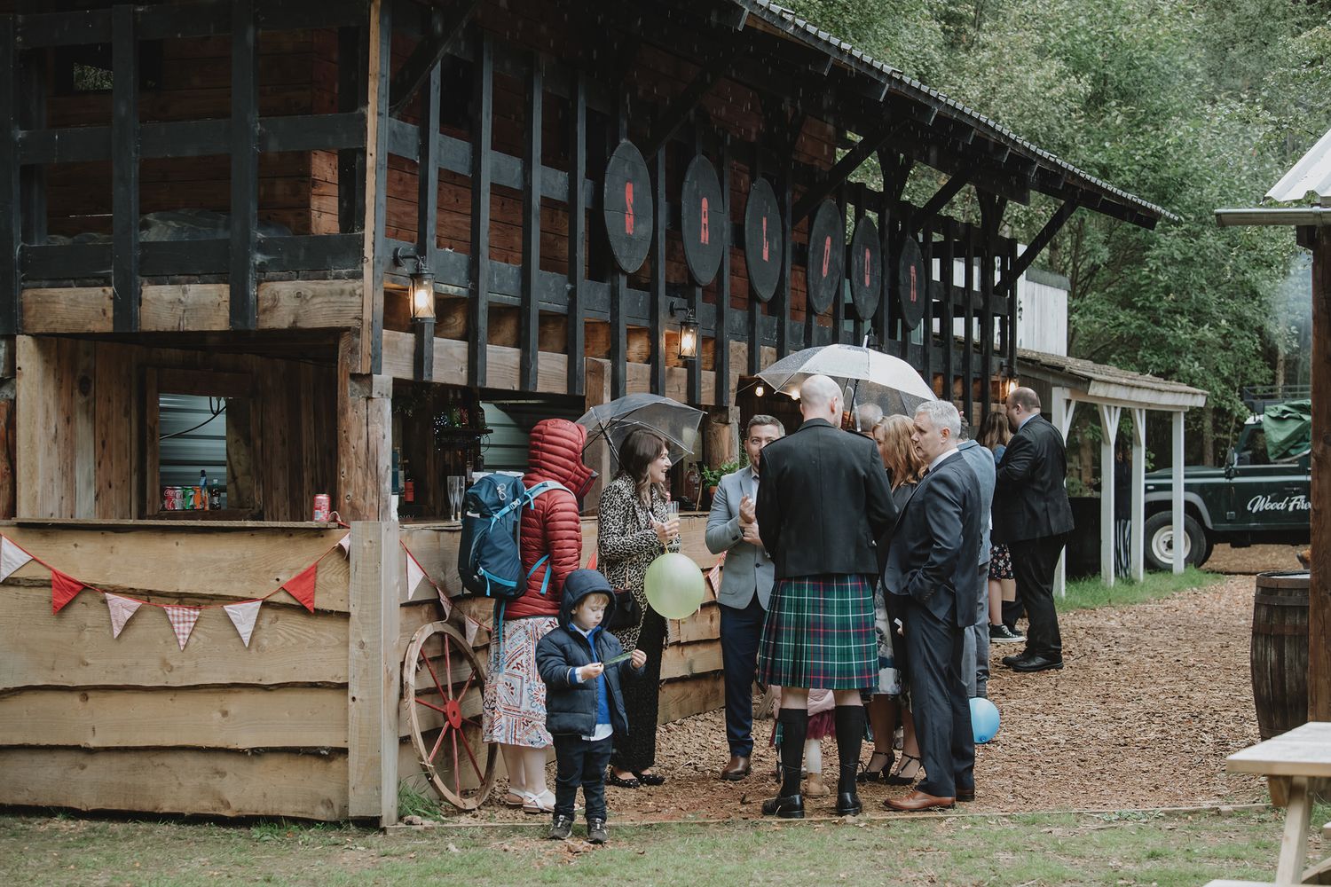 Wedding guests with umbrellas gather outside a rustic wooden venue in kilts.