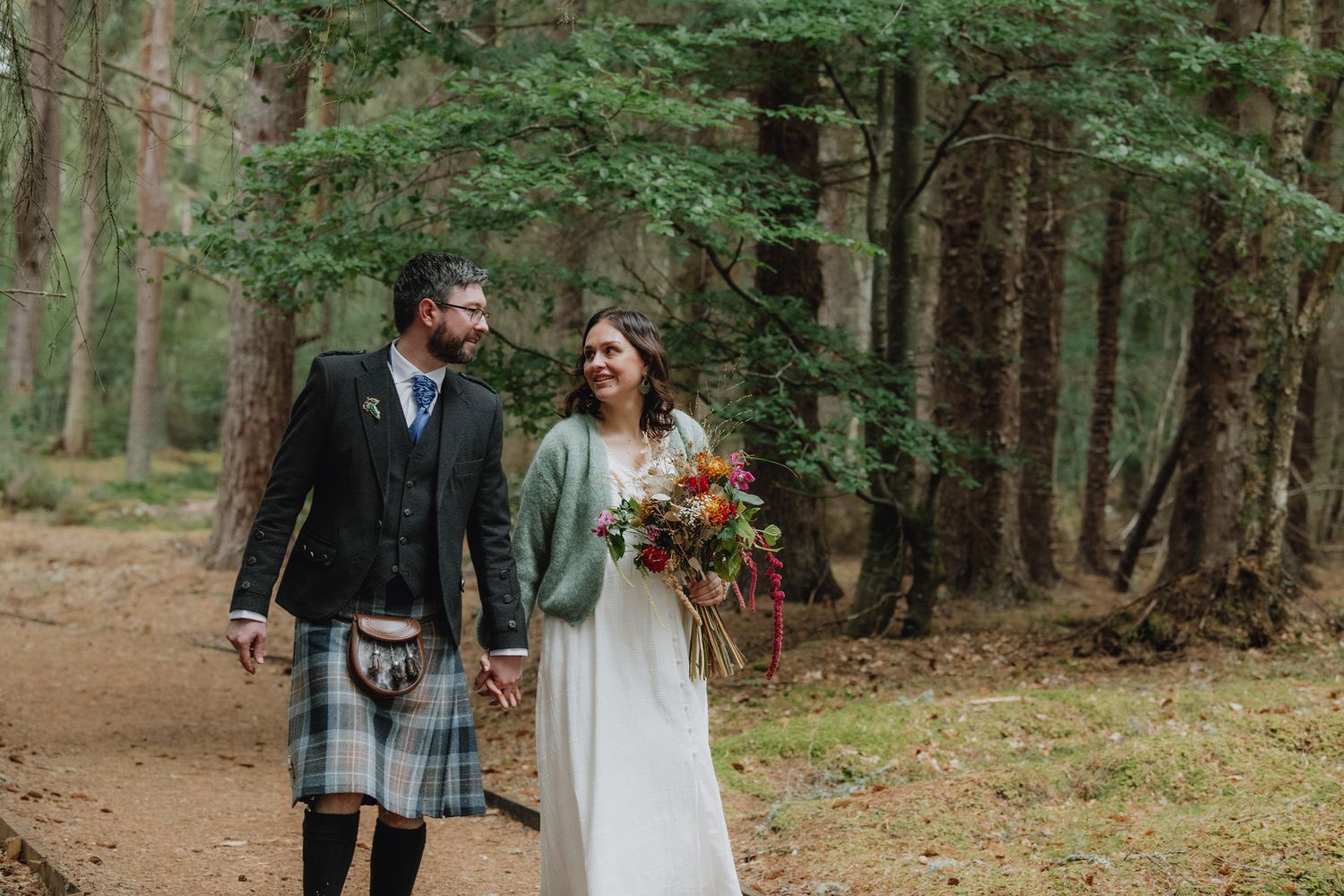 Bride and groom walking hand in hand through a pine forest, groom wearing traditional Scottish tartan kilt.