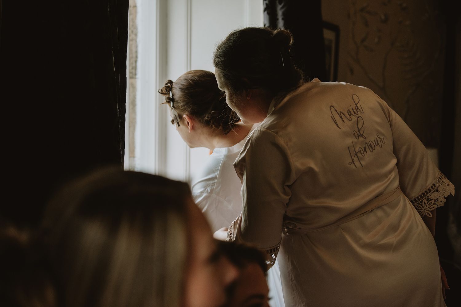 Bride in satin robe with flower girl looking out window during wedding morning preparations.