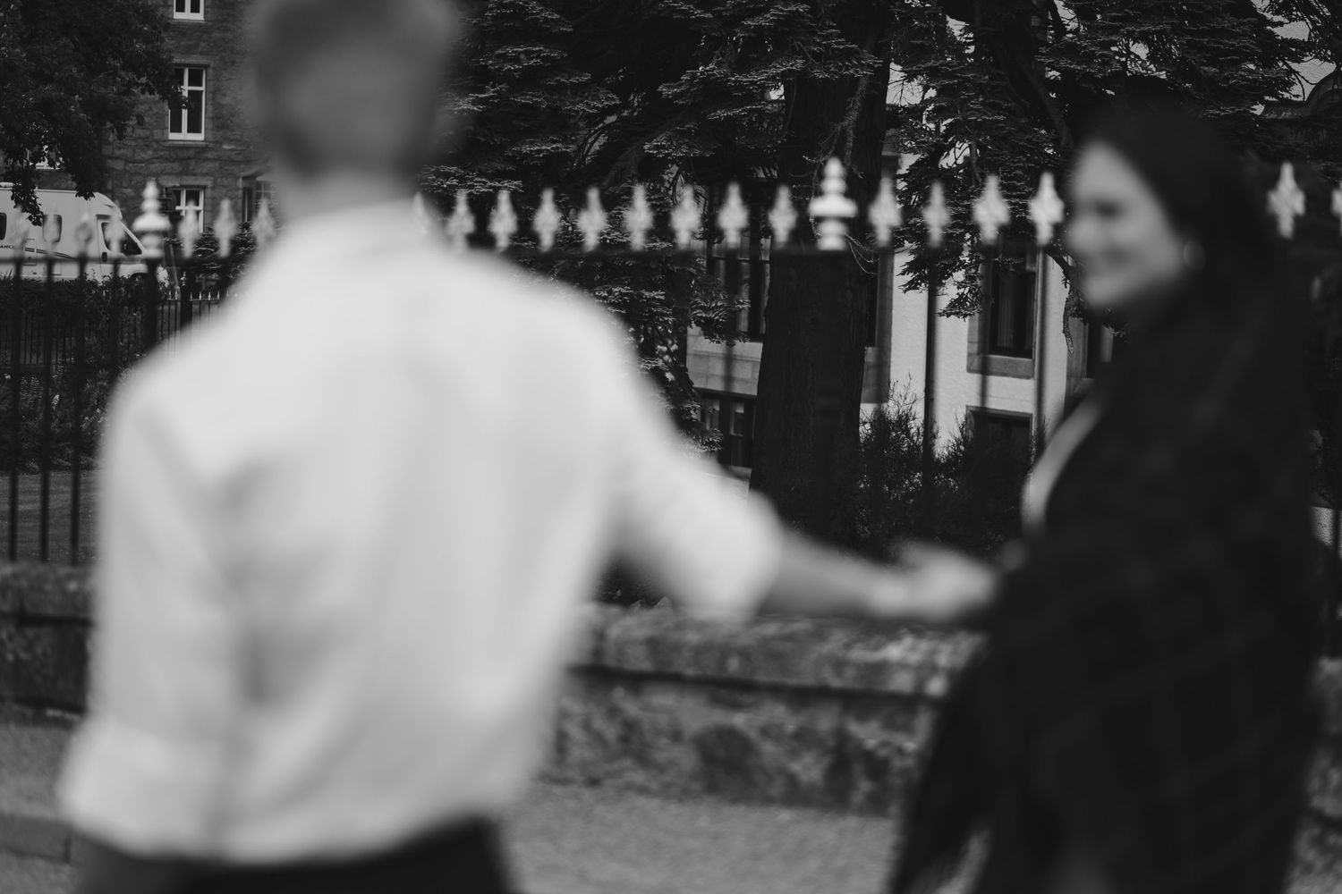 Couple holding hands near an ornate white fence in a soft-focus romantic outdoor setting.