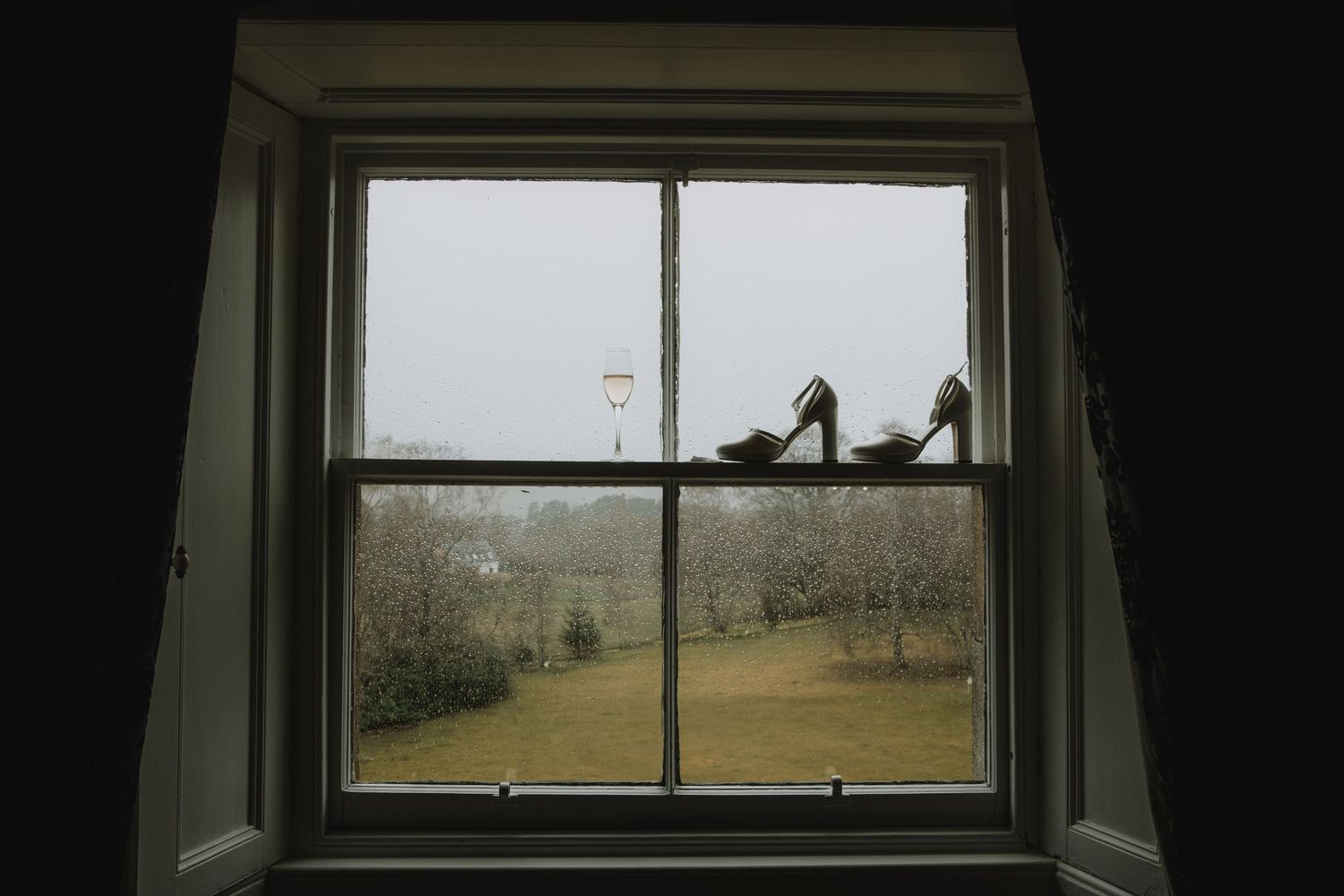 Elegant bridal heels silhouetted on windowsill with countryside view on a misty wedding morning.