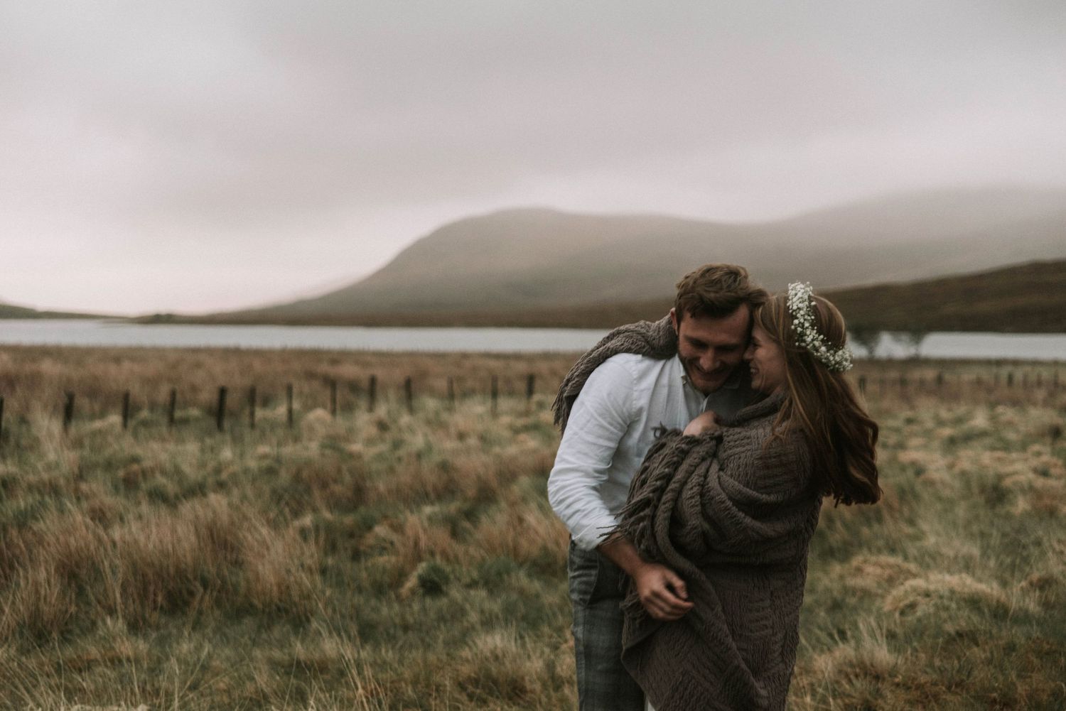 Couple laughing and embracing in grassy Scottish Highland field beside a misty loch.