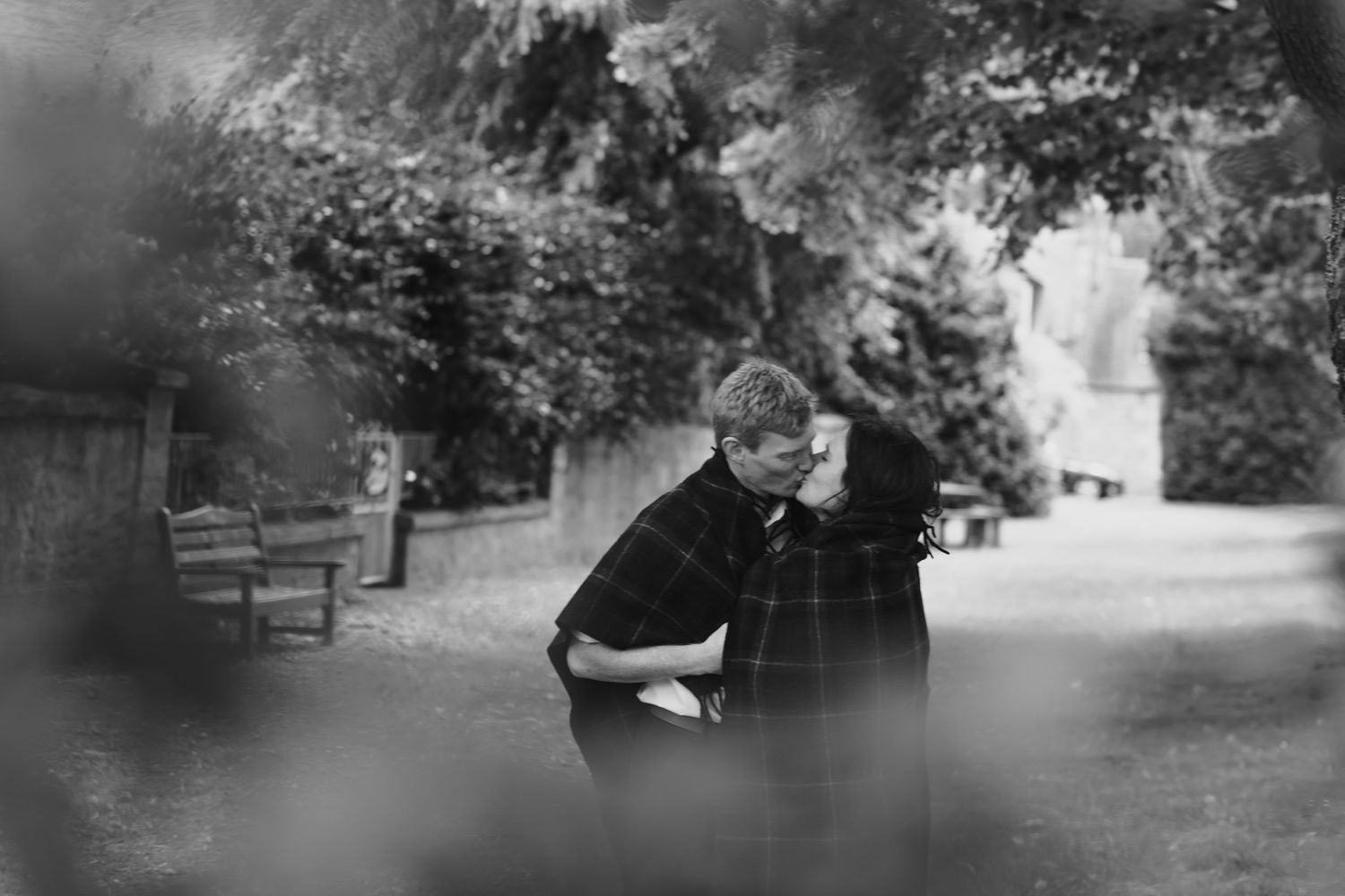 Black and white photo of a couple embracing in a lush park setting with benches and trees in background.