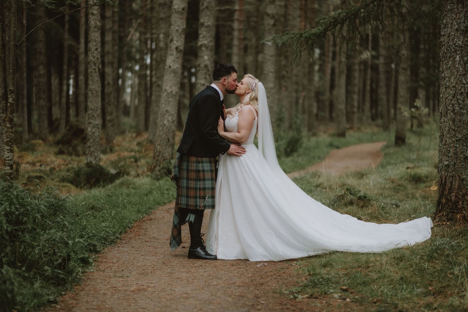 Bride and groom sharing a romantic kiss on a forest path, groom wearing a traditional Scottish tartan kilt.
