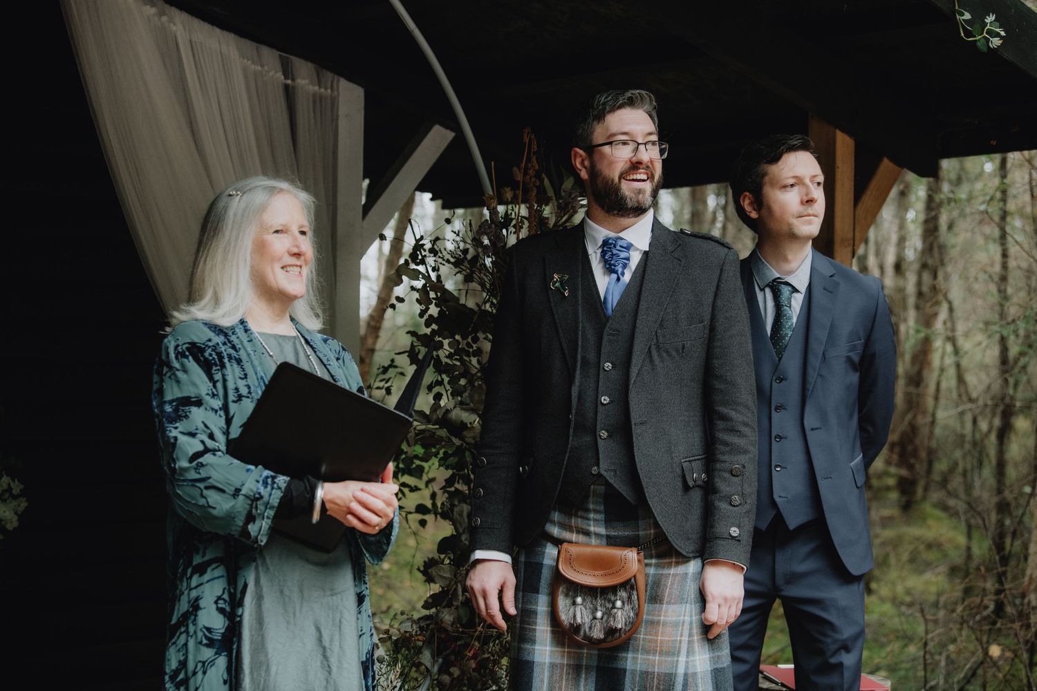 Groom in tartan kilt standing with officiant and groomsman under a woodland ceremony canopy.
