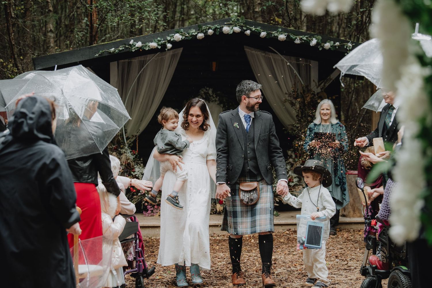 Newlywed couple walks down forest aisle with children as guests hold umbrellas at rainy woodland wedding.