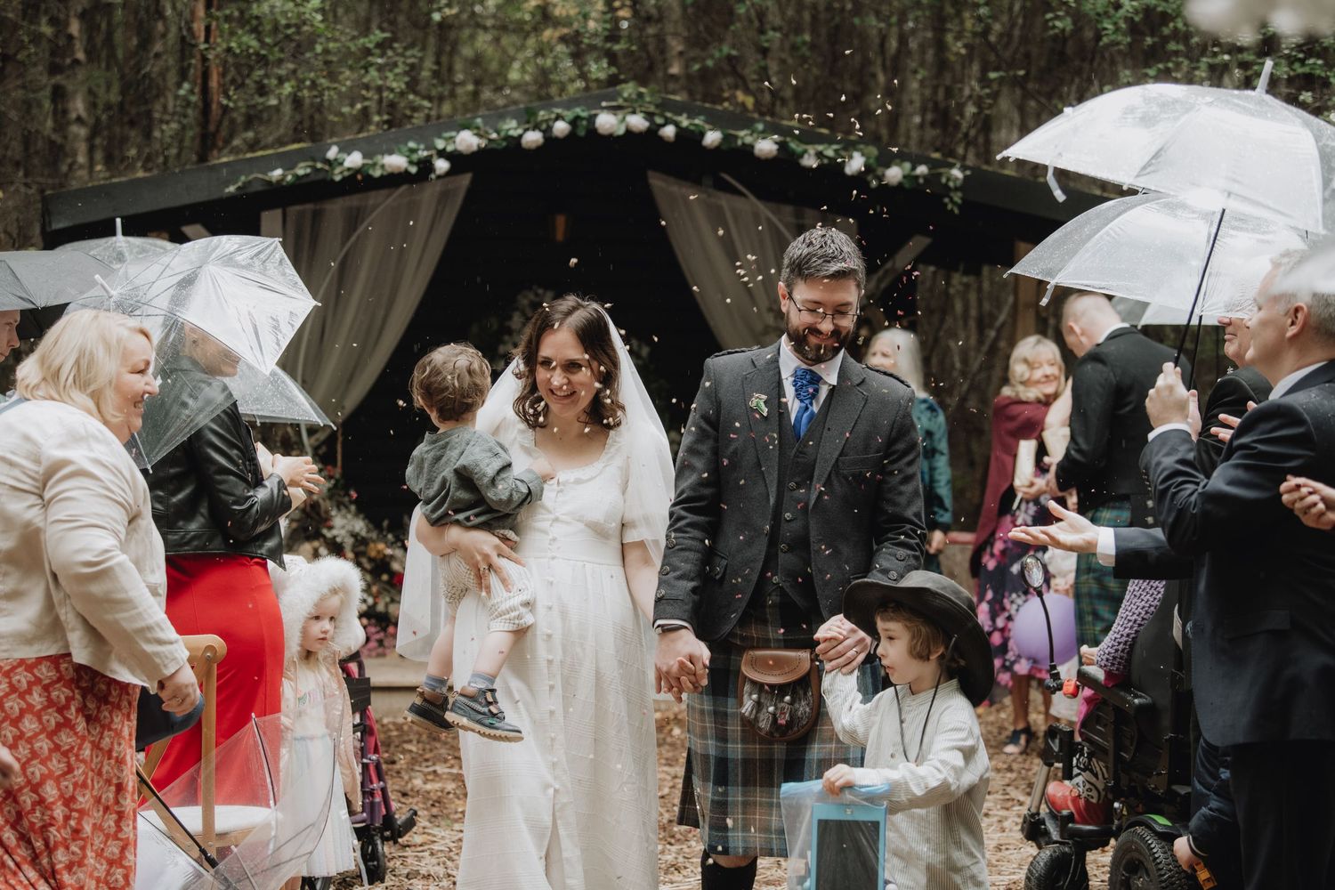 Happy couple exits woodland wedding ceremony with children as guests celebrate with umbrellas in forest.