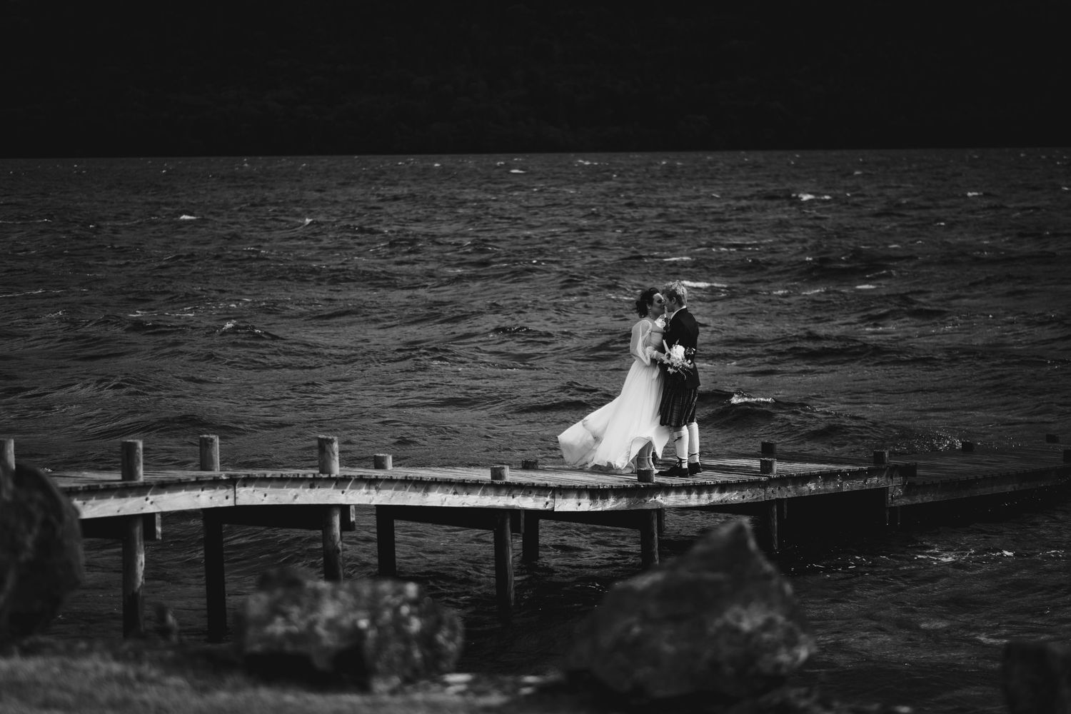 Black and white photo of bride and groom embracing on a wooden pier beside a dramatic windswept loch.