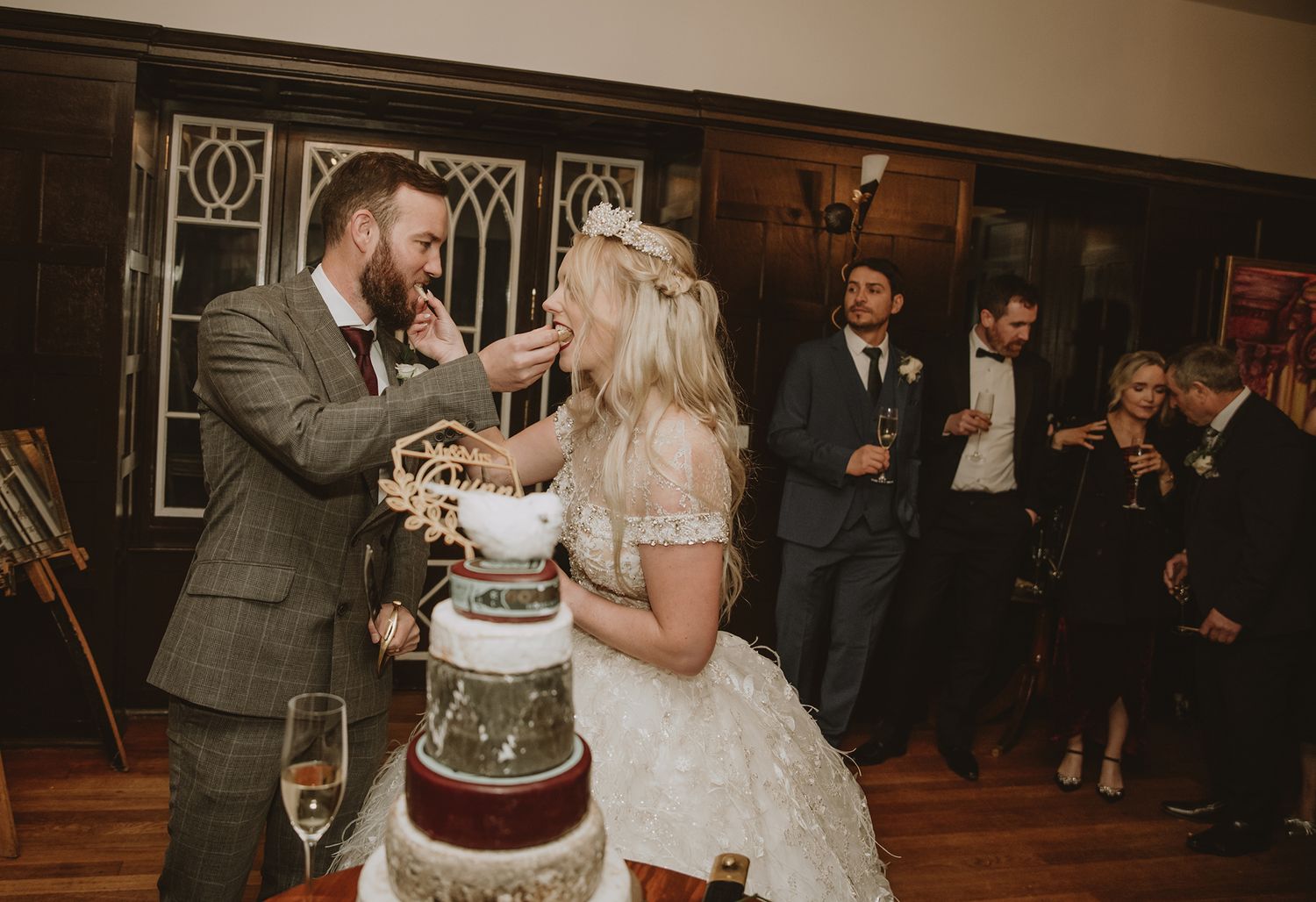 Bride and groom cutting wedding cake together at elegant indoor reception while guests celebrate in background.