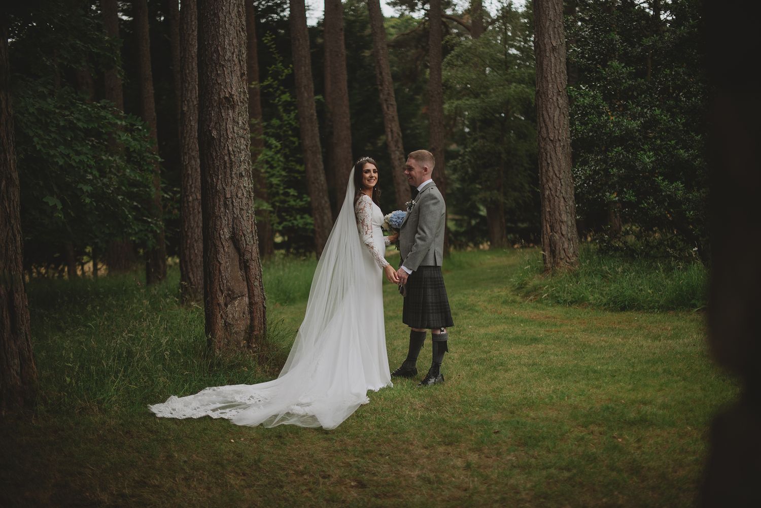 Bride in white gown with veil and groom in Scottish kilt standing together in a lush forest setting.