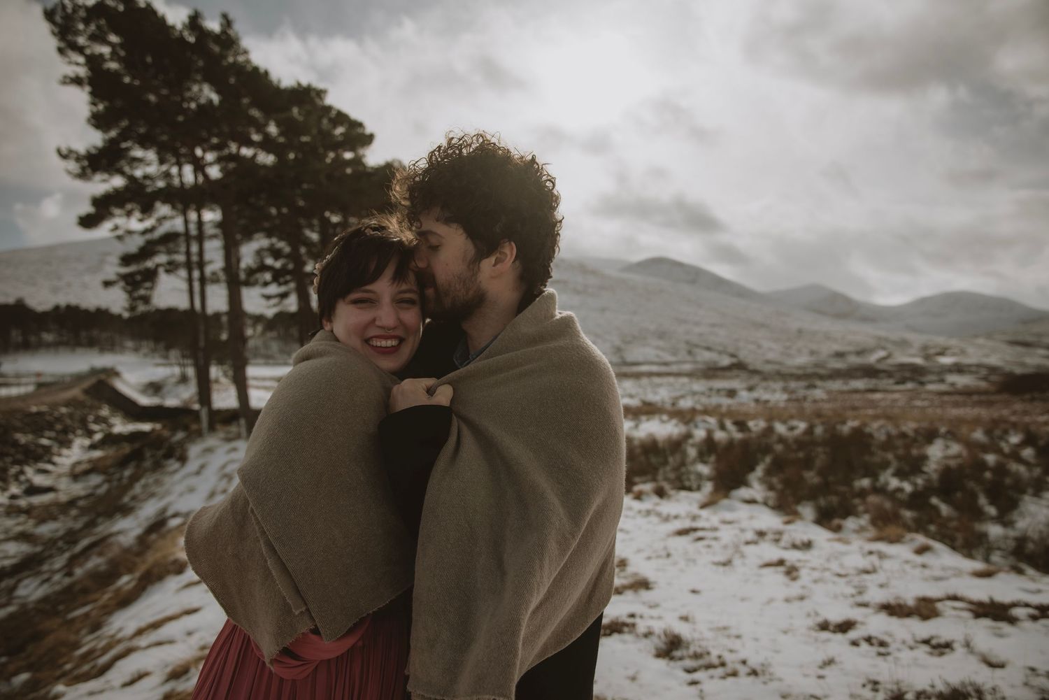 Couple wrapped in blanket sharing romantic embrace in snowy Scottish Highland landscape with mountains in background.
