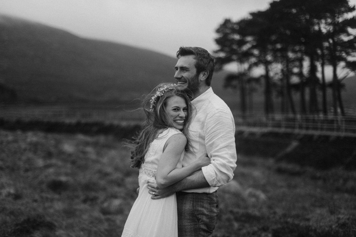 Black and white wedding portrait of smiling bride and groom embracing in a highland landscape.