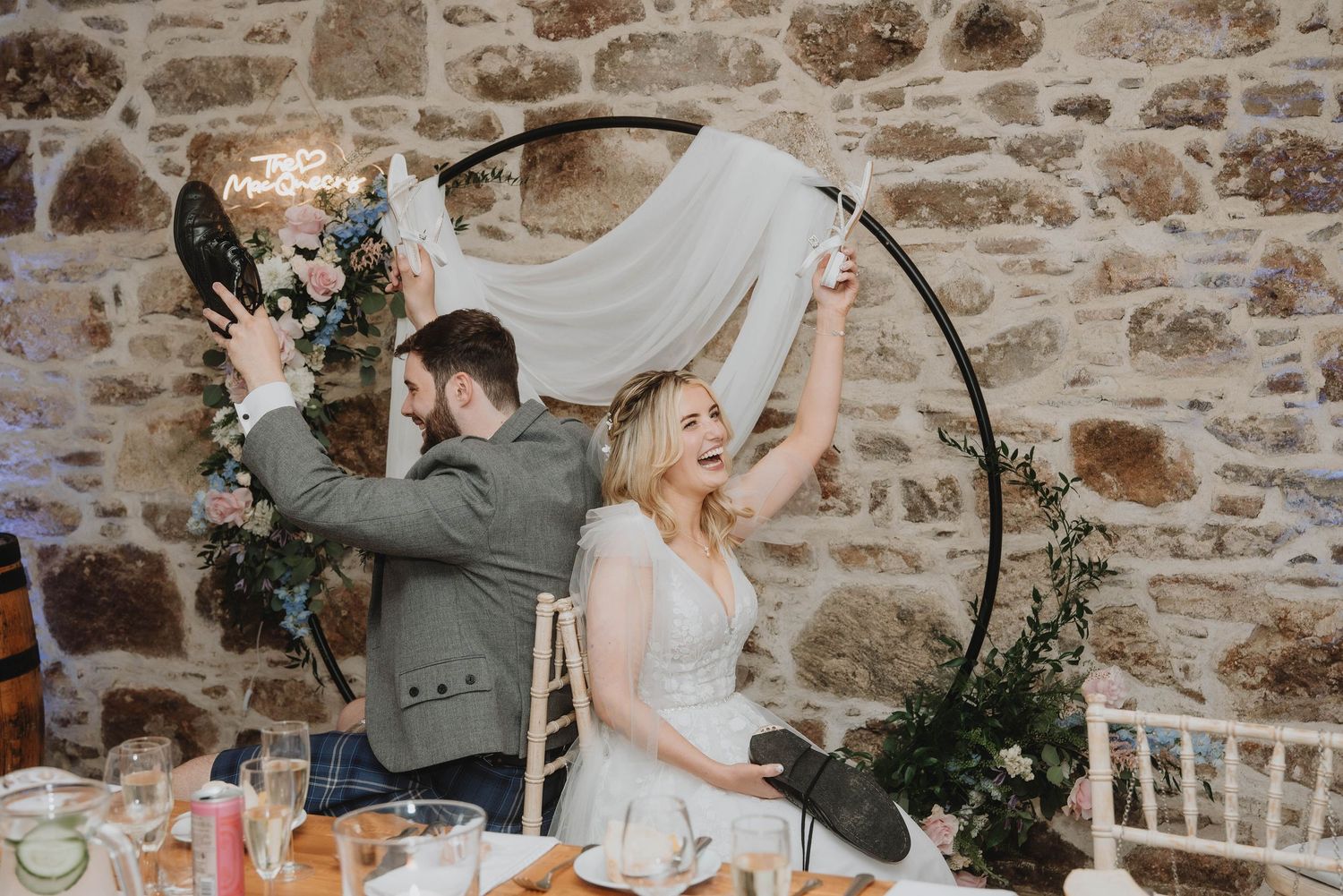 Bride and groom celebrating at wedding reception table in front of rustic stone wall with floral hoop.