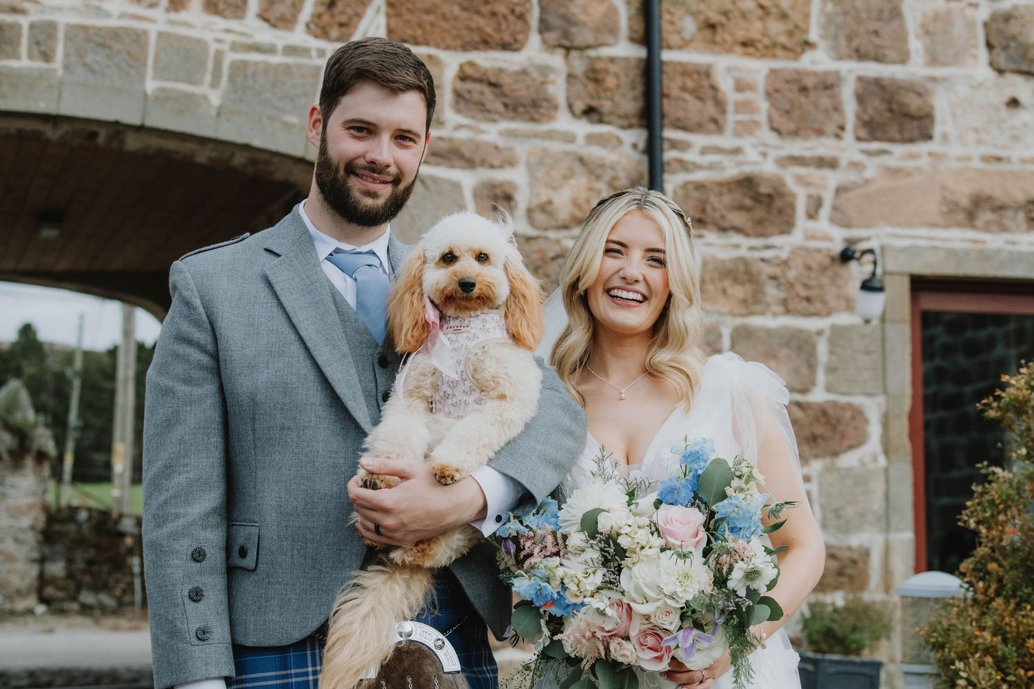 Happy bride and groom pose with their golden doodle dog outside rustic stone wedding venue.