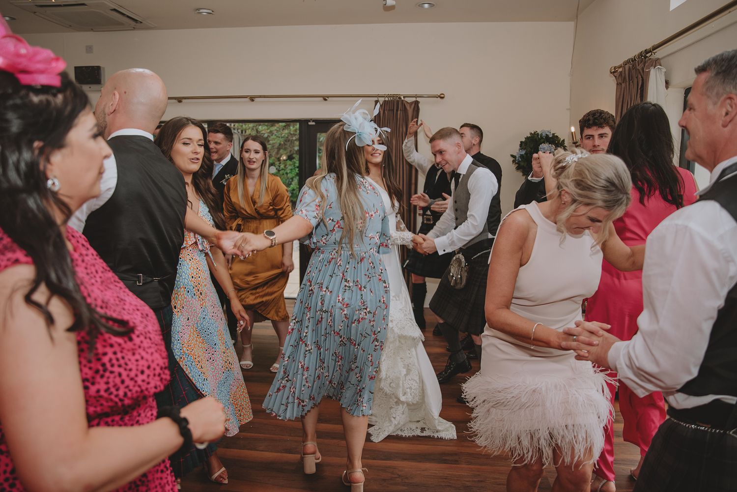 Wedding guests dancing in a circle at a Scottish wedding reception, with bride in white and guests in colorful formal attire.