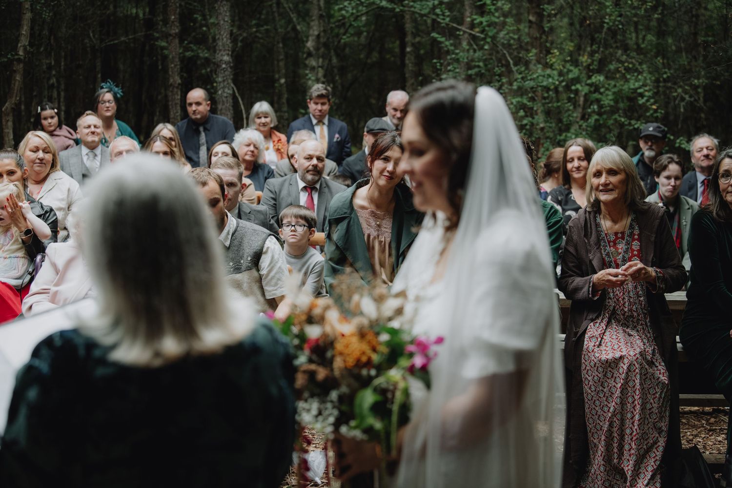 Bride walks down forest aisle as emotional wedding guests applaud and smile during outdoor woodland ceremony.