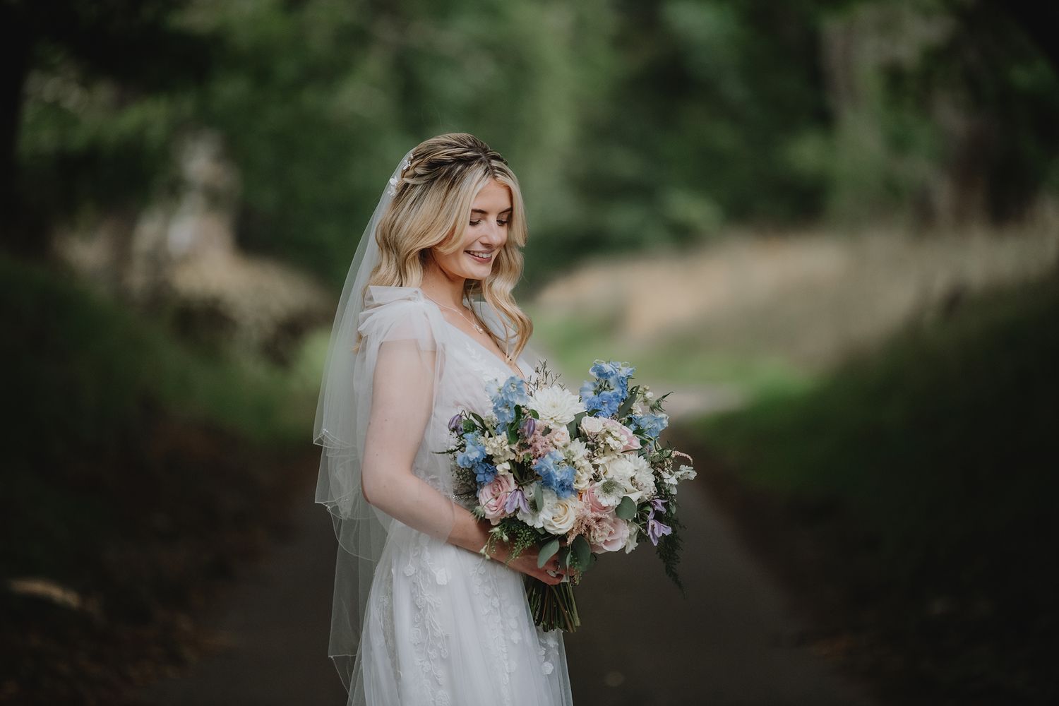 Smiling bride holding blue and white floral bouquet wearing veil and floral crown in green woodland.