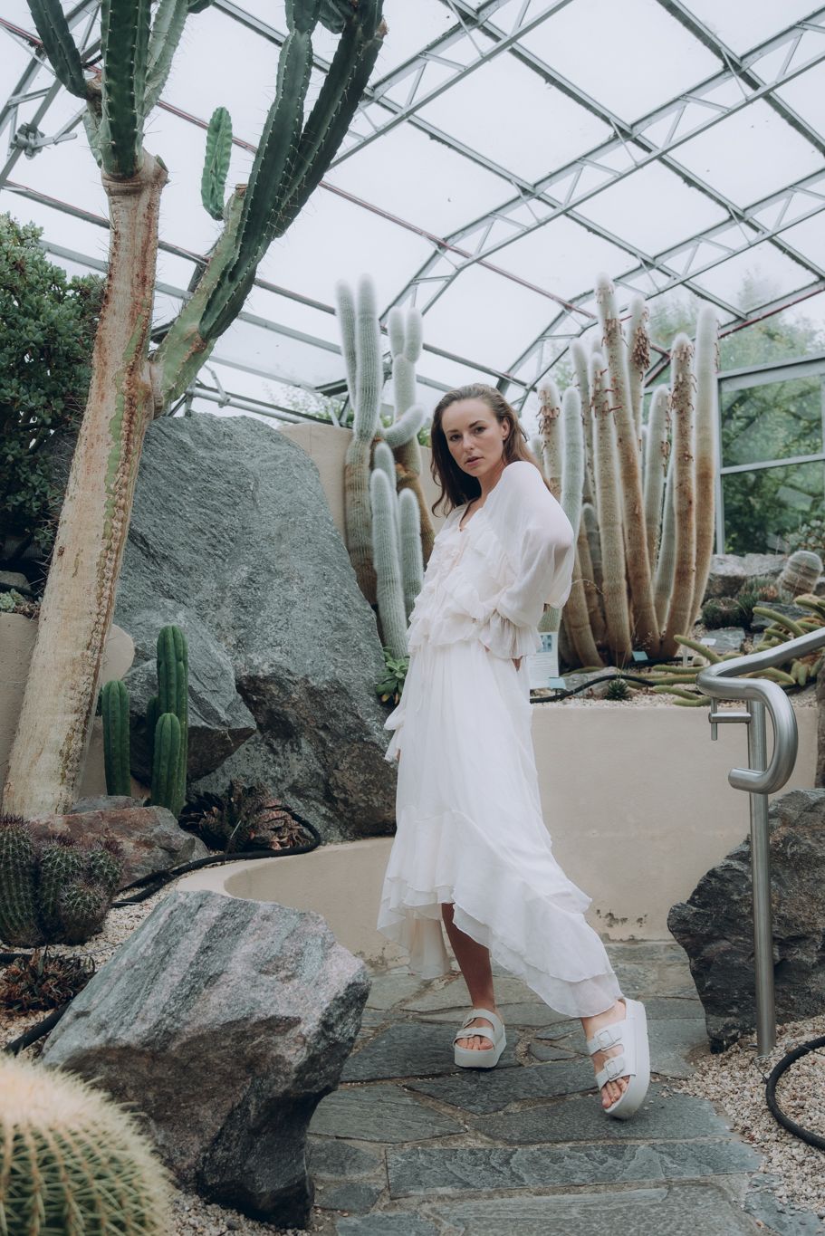 Woman in white ruffled dress and slide sandals posing among large cacti in a glass greenhouse.