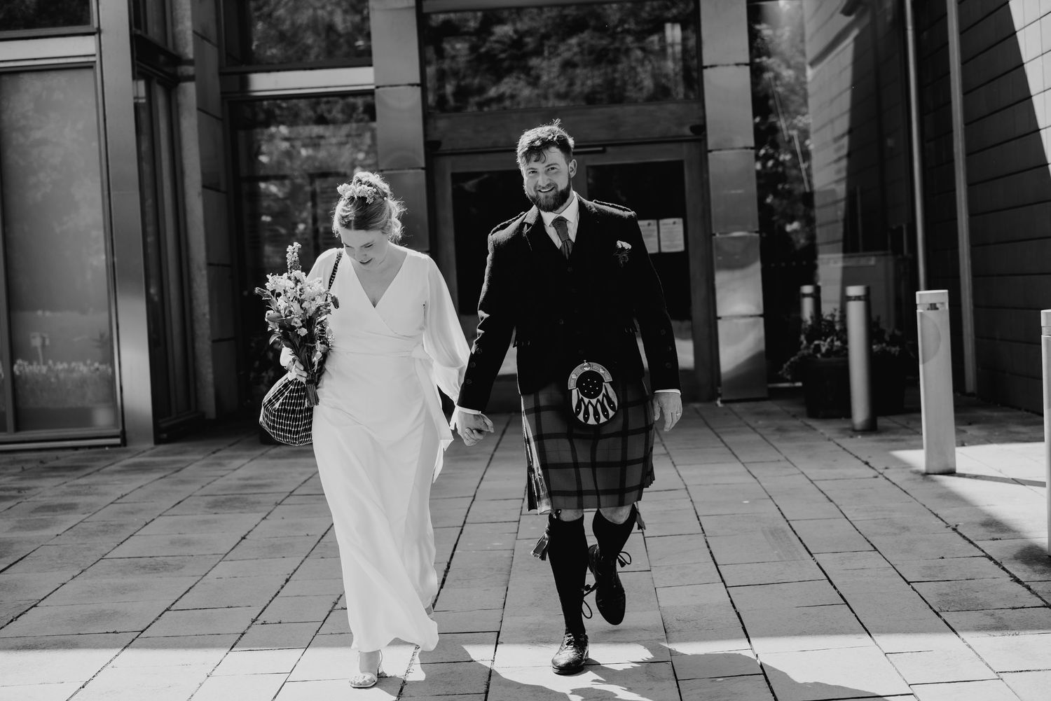 Black and white photo of newlyweds walking hand in hand, groom wearing traditional Scottish kilt.