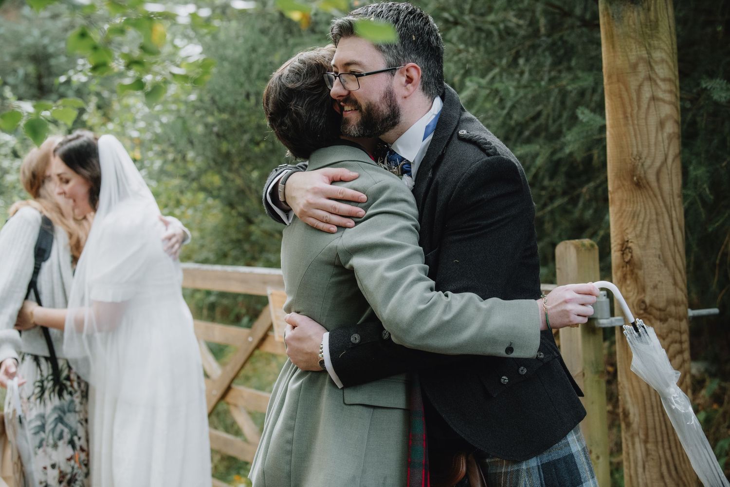 Groom in tartan kilt shares warm embrace with male guest at outdoor woodland wedding celebration.