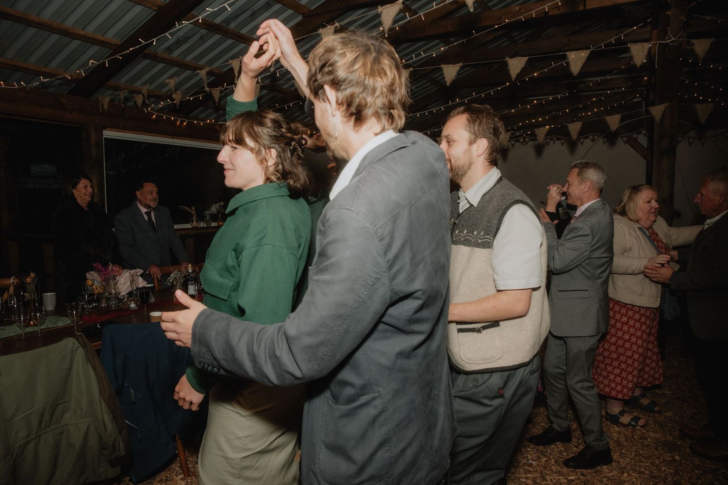 Couple dancing together at a rustic wedding reception with bunting decorations and guests in background.