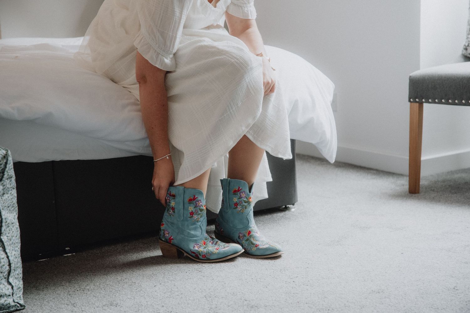 Bride in white wedding dress wearing embroidered turquoise cowboy boots sitting on edge of bed.
