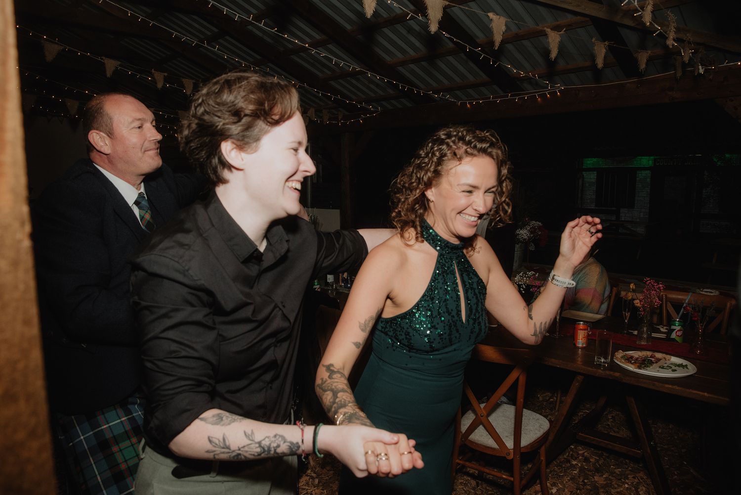 Two women laughing and dancing joyfully at a wedding reception in a rustic barn venue with string lights.