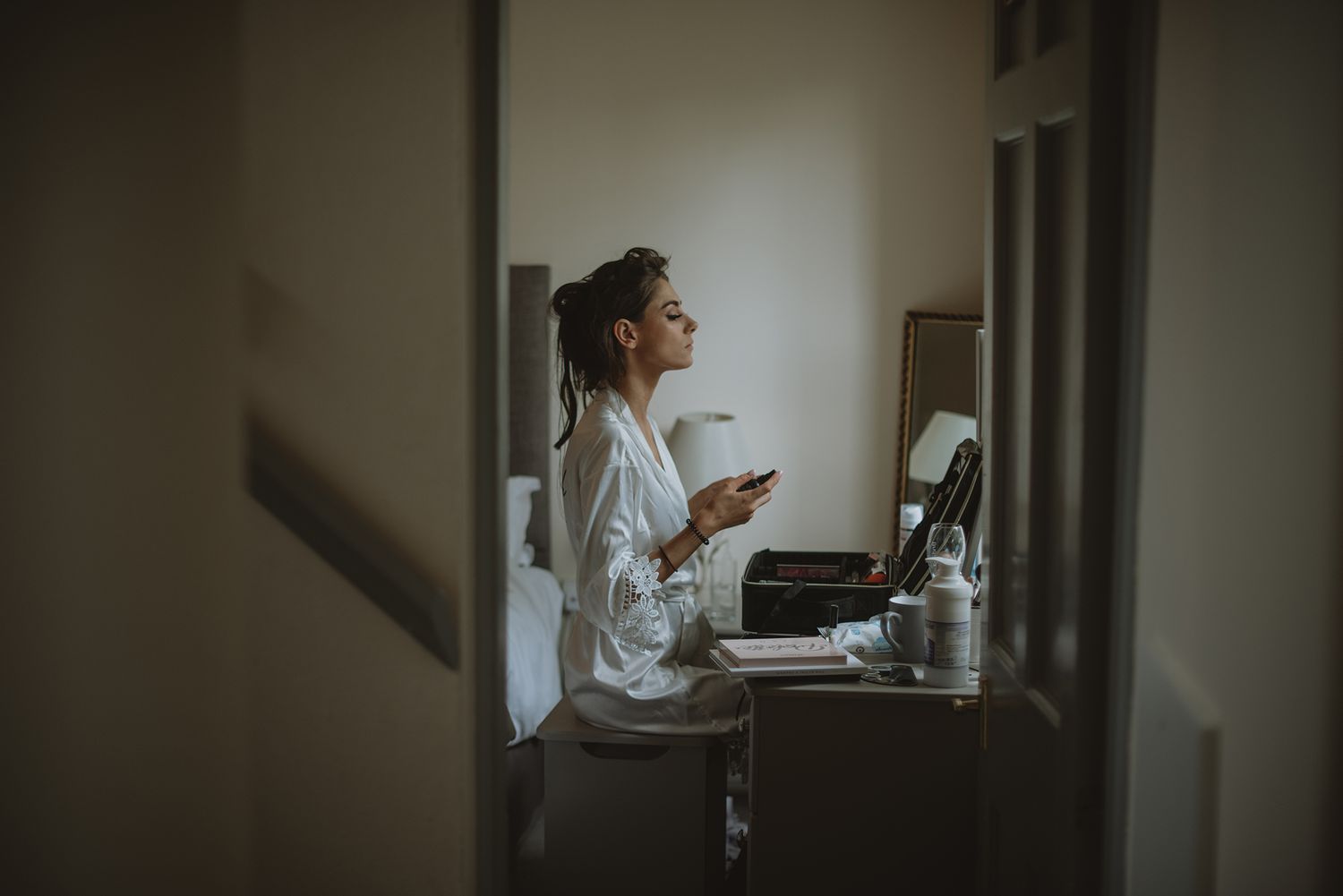 Bride in white robe sits at vanity mirror applying makeup in a softly lit hotel room.