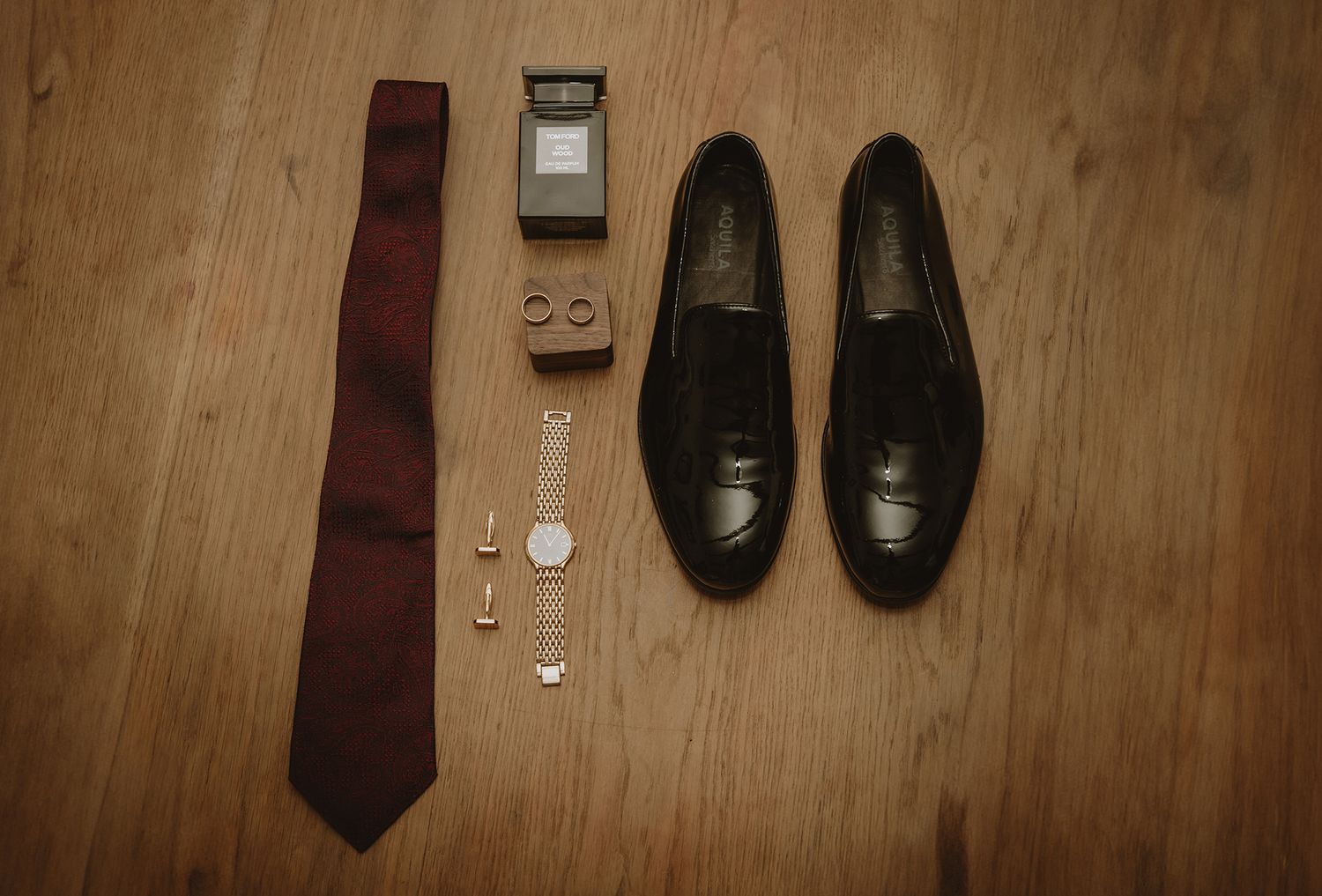 Flat lay of men's wedding accessories including burgundy tie, black loafers, cufflinks, cologne, and gold watch on wood floor.
