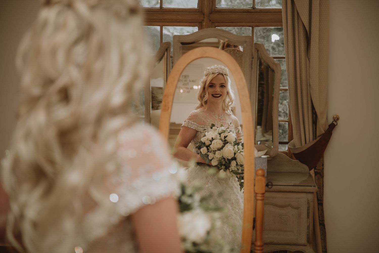 Bride with blonde hair admires herself in an ornate mirror, holding a white bouquet in her lace wedding dress.