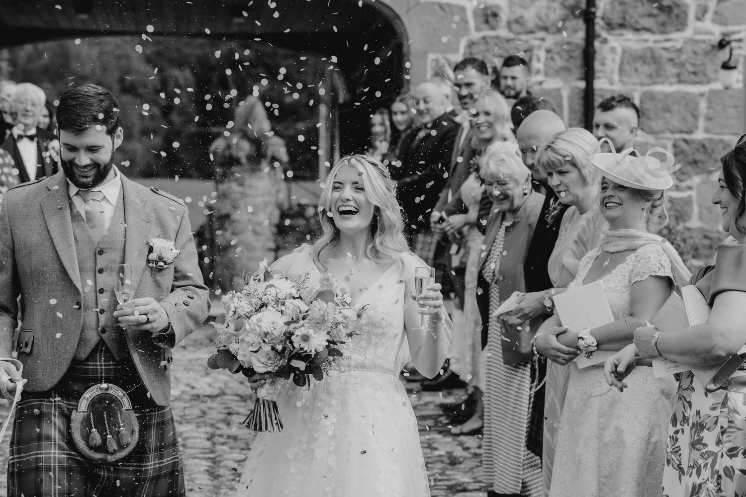 Black and white photo of bride laughing joyfully as guests throw confetti outside wedding venue.