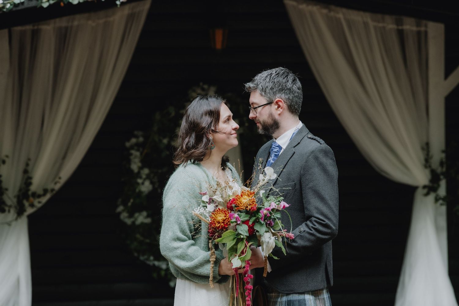 Bride and groom share an intimate moment under draped fabric arch, bride holding vibrant autumn bouquet.