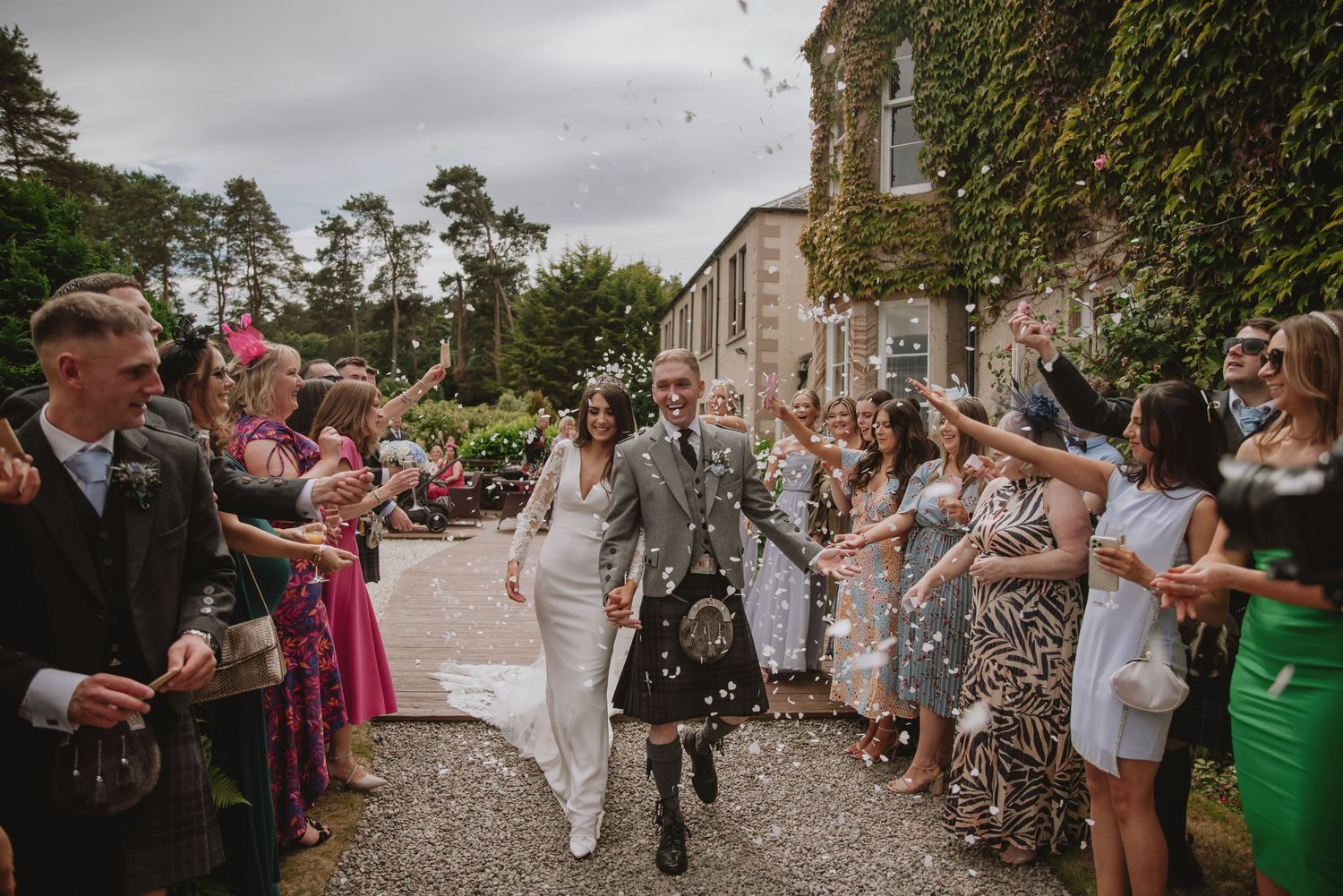 Bride and groom walk through confetti shower outside ivy-covered Scottish castle venue surrounded by cheering wedding guests.