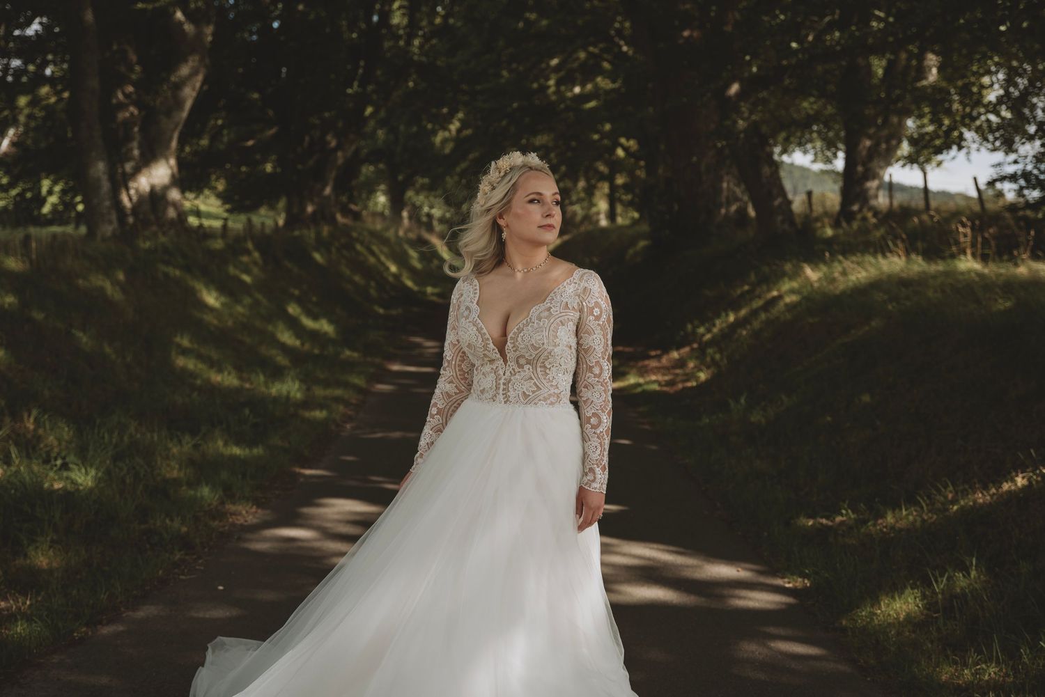 Bride in lace long-sleeve wedding gown posing on a sunlit tree-lined path.